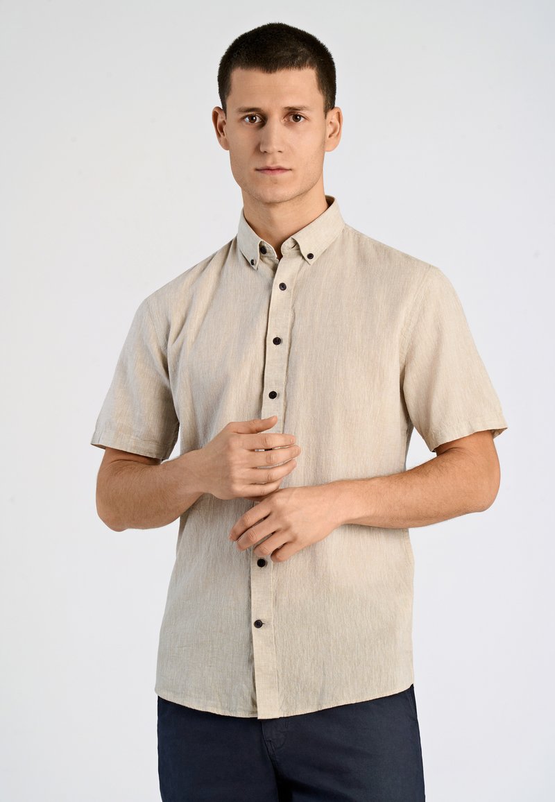 Camisa de botones de manga corta en color beige, confeccionada en una tela texturizada. Presenta un cuello puntiagudo y botones oscuros, combinada con pantalones oscuros.