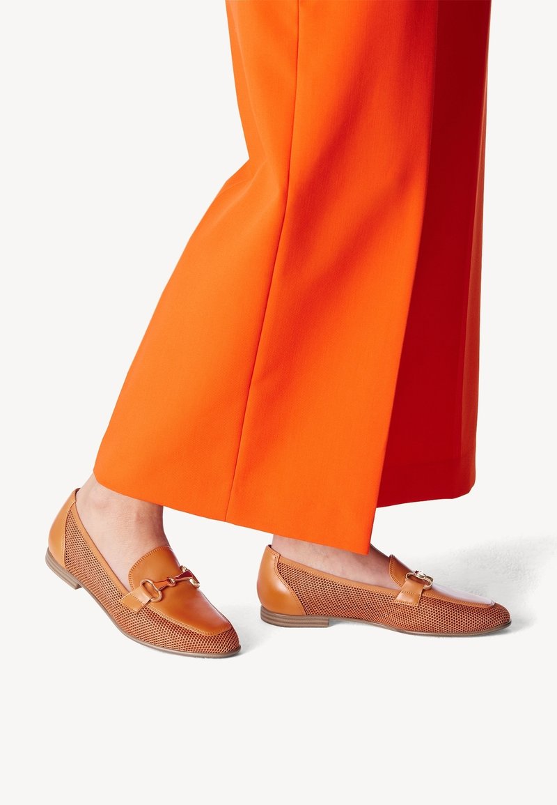 Tan leren loafers met geperforeerde textuur en platte zolen, met een metalen accent. Gepaarde met feloranje wijde broekspijpen.
