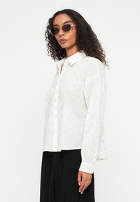 Biała koszula zapinana na guziki z metalicznym wzorem kropek, długie rękawy i kołnierzyk. Modelka nosi ją z czarnymi spodniami oraz dużymi okularami przeciwsłonecznymi.