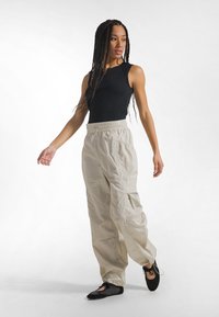 Kvinde med langt fletninghår iført en sort ærmeløs top, beige cargobukser og sorte balletflade sko, stående på en hvid baggrund.