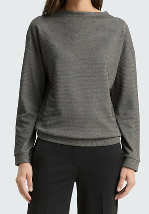 Donna che indossa una maglia aderente a maniche lunghe con motivo bianco e nero e scollo a barca, pantaloni neri, in piedi con le braccia rilassate.