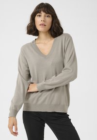 Hellbrauner V-Ausschnitt-Pullover mit langen Ärmeln, gerippten Bündchen und Saum. Glatte Textur, lässige Passform, kombiniert mit schwarzen Hosen.