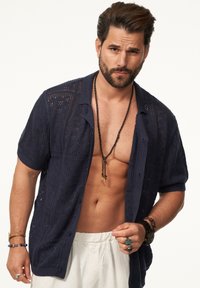 Navyblaues Strickshirt mit kurzen Ärmeln und strukturiertem offenen Muster, kombiniert mit cremefarbenen Hosen, verziert mit Perlen und geschichteten Halsketten.