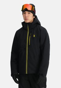 Schwarze Skijacke mit gelben Reißverschlussakzenten, wasserdichtem Material, hohem Kragen und abnehmbarer Kapuze. Getragen mit schwarzen Handschuhen und Skibrille.