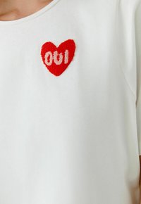 Witte T-shirt met korte mouwen en een rood geborduurd hart met de tekst "001" in lichtroze, geplaatst op de linkerborst.