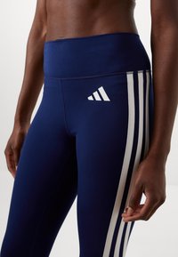 Marineblauwe leggings van rekbaar materiaal met een hoge tailleband, voorzien van drie witte strepen langs de zijkant en een Adidas-logo.
