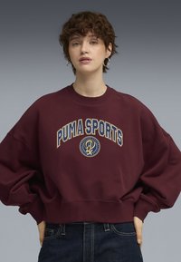 Burgunder sweatshirt med løs pasform, ribbet halsudskæring og oversized ærmer. Har "PUMA SPORTS" grafik i creme og guld.