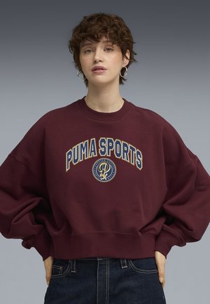 Burgunder sweatshirt med løs pasform, ribbet halsudskæring og oversized ærmer. Har "PUMA SPORTS" grafik i creme og guld.
