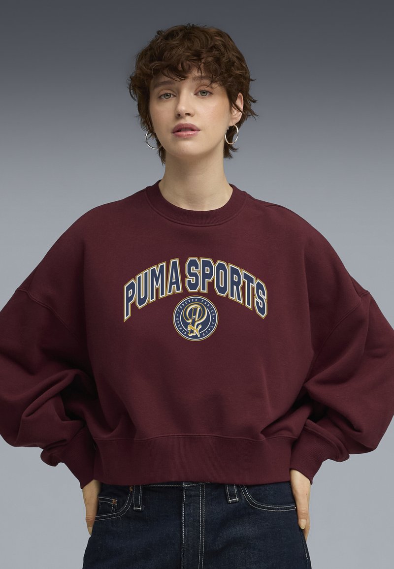 Burgunder sweatshirt med løs pasform, ribbet halsudskæring og oversized ærmer. Har "PUMA SPORTS" grafik i creme og guld.