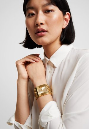 Montre en or avec un cadran rectangulaire et un bracelet en maillons, portée sur une chemise blanche. Surface lisse, finition polie et design minimaliste.