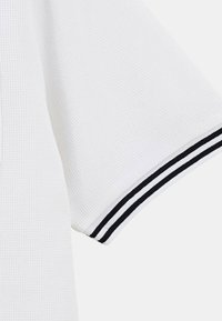 Polo shirt blanc avec un tissu texturé, des manches courtes et des accents rayés noir et blanc sur les poignets.