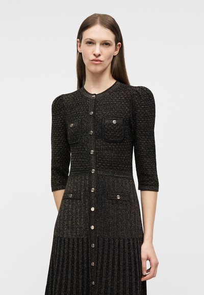 Γυναίκα με ίσια καστανά μαλλιά φοράει μαύρο υφασμάτινο μεσαίο φόρεμα με φουσκωτούς ώμους, εμπρός με κουμπιά και τσέπες στο στήθος και στους γοφούς.