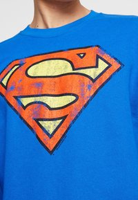 T-shirt di cotone blu con un grande logo di Superman sbiadito in rosso e giallo. Collo a giro e maniche corte, con una stampa grafica testurizzata.