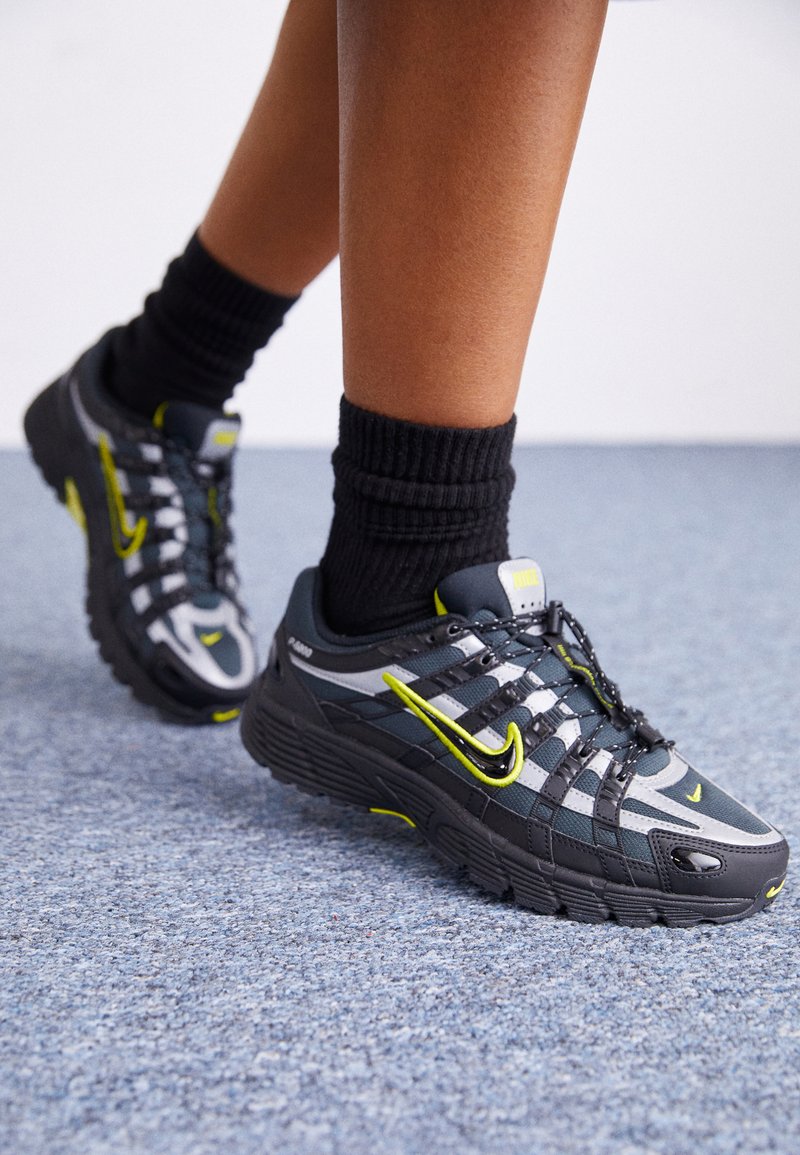 Zapatillas deportivas en negro y gris con acentos amarillos, que cuentan con una parte superior de malla, suela gruesa y un sistema de cordones prominente.