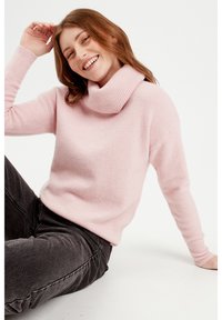 Pull en tricot rose pâle avec un col roulé replié et côtelé, des manches longues et une coupe décontractée. Associé avec un jean gris foncé.