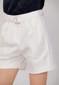 Shorts blancs sur mesure en tissu lisse, dotés de plis et d'une ceinture avec boucle circulaire en argent. Coupe légèrement ample avec un ourlet soigné.