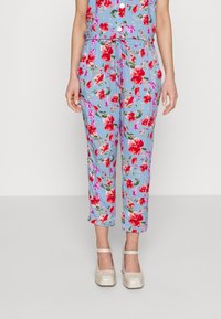 Femme portant un pantalon cropped floral bleu avec des fleurs rouges et roses, associé à des escarpins de style Mary Jane beige.