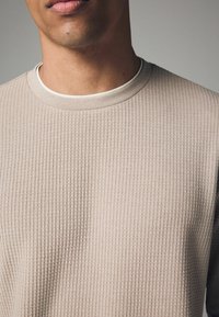 Sudadera beige texturizada con un patrón acanalado y un cuello redondo, que presenta un collar interior más claro y un diseño de corte holgado.
