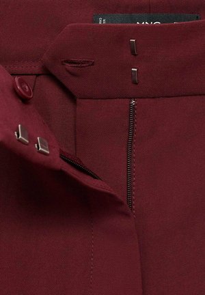 Pantalons marron avec fermeture éclair, bouton et deux attaches métalliques. Le tissu a une texture lisse avec des détails de couture.