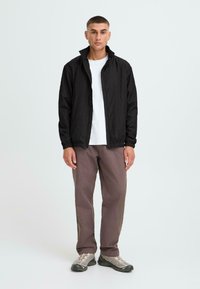 Black lightweight jacket with a zip and elastic cuffs, worn over a white t-shirt; paired with brown trousers and gray shoes.