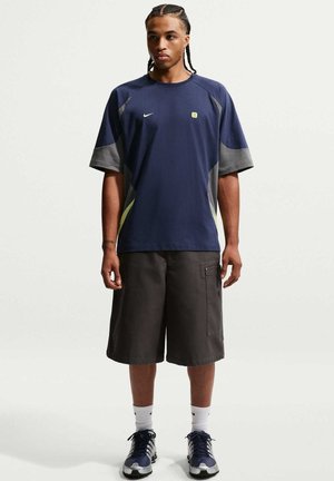 Persoon staat naar voren gericht, draagt een marineblauw T-shirt, lange donkere shorts, witte sokken en marineblauwe sneakers tegen een effen achtergrond.