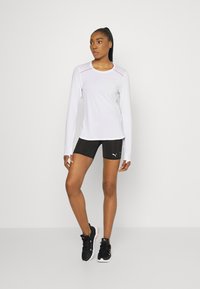 Valge pika varrukaga sportlik topp roosa detailidega, kombineeritud mustade lühikeste pükstega. Modell kannab musti spordijalatu, seistes neutraalses taustas.