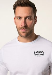 Camiseta blanca de manga larga con cuello redondo acanalado y texto impreso en negro "BANDERA RODEO TEXAS EST. 1978" en el lado izquierdo del pecho. Tela de algodón suave.
