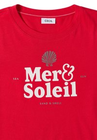 T-shirt rouge avec texte blanc et gris "Mer & Soleil", petites inscriptions "Sea", "Sun" et "Sand & Shell", ainsi qu'un motif de coquillage gris au-dessus du texte.