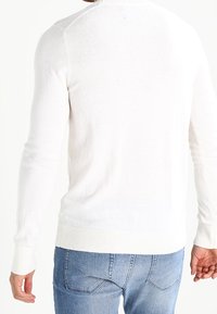 Suéter de punto blanco con mangas largas, puños y dobladillo acanalados, y cuello redondo. Se lleva con vaqueros azules, mostrando una textura suave y un diseño ligero.