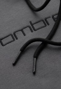 Zbliżenie czarnych sznurków ściągających z markowymi plastikowymi końcówkami spoczywających na ciemnoszarej tkaninie wyszytej fragmentem logo "Ombre".