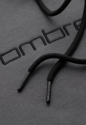 Makro záběr na černé šňůrky s označením a plastovými špičkami odpočívajícími na tmavě šedé látce vyšívané částí loga "Ombre".