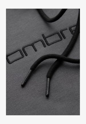 Makro záběr na černé šňůrky s označením a plastovými špičkami odpočívajícími na tmavě šedé látce vyšívané částí loga "Ombre".