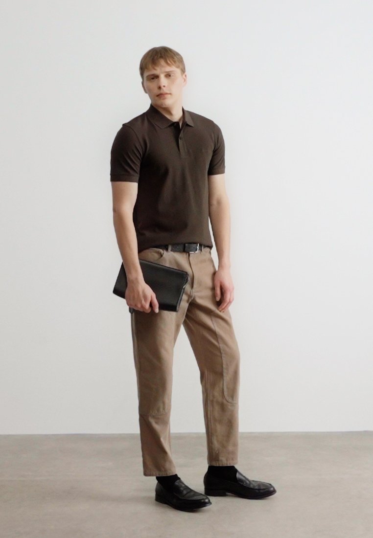 Jeune homme debout, portant un polo marron foncé, un pantalon beige, une ceinture noire, des chaussures noires, et tenant une pochette noire dans sa main gauche.