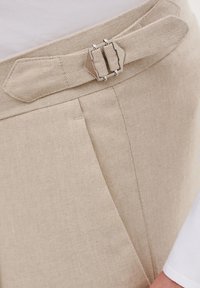 Beżowy pasek z tkaniny z prostokątną metalową klamrą, przymocowany do spodni z fałdami z przodu i gładką teksturą.