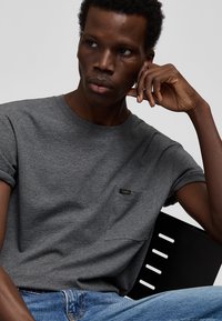 Nuori tummaihoinen mies, jolla on lyhyet kiharat hiukset, pukeutunut harmaaseen Lee-t-paitaan ja sinisiin farkkuihin, istuu käden poskella.