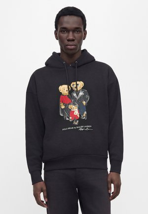 Homme portant un sweat à capuche noir avec un graphisme de trois ours en peluche habillés et le texte Polo Bear de Ralph Lauren.