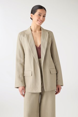 Donna sorridente, indossa un blazer beige e pantaloni a gamba larga abbinati sopra una maglia color prugna intenso, in piedi davanti a uno sfondo chiaro e uniforme.
