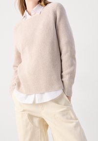 Pull tricoté beige clair à texture côtelée, col rond et manches longues, superposé sur une chemise blanche à col.