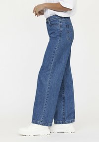Jeans de denim azul de pierna ancha con una cintura alta, una sutil textura deslavada y costuras en contraste. Combinados con zapatillas deportivas blancas.