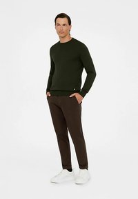 Tamsiai žalias megztinis su apvalia iškirpte, suknelės rankogaliai, derinamas su rudos spalvos kelnėmis ir baltais sportbačiais, stovintis prieš paprastą fono.