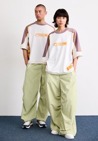 Witte sportieve t-shirts met paarse accenten en een oranje logo, gecombineerd met lichtgroene loszittende broek. Beiden dragen sportieve sneakers.