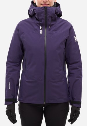 Person trägt eine lila wasserdichte Jacke mit Kapuze, schwarze Handschuhe und schwarze Hose, steht vor einem einfarbigen Hintergrund.