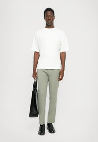 White cotton t-shirt, loose fit, short sleeves. Light gray checkered trousers, tailored fit. Model holds a black leather tote bag. Black shoes.