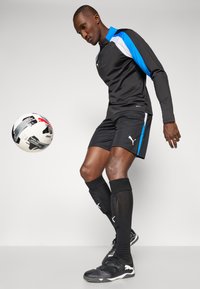 Svart sportsklær bestående av langermet topp med blå detaljer, svarte shorts og svarte knelange sokker. En svart og hvit fotball ligger i nærheten.