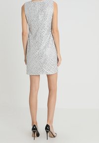 Robe sans manches argentée avec un motif texturé en sequins, dotée d'une fermeture à glissière au dos et associée à des chaussures à talons hauts noires.