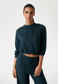 Gekürzte Kapuzenjacke in dunklem Türkis, aus leichtem Stoff. Mit einem Frontreißverschluss, Logodetail und elastischem Saum. Kombiniert mit passenden Hosen.