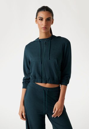 Cropped hooded jacket in dark teal, made of lightweight fabric. Features a front zipper, logo detail, and elasticized hem. Paired with matching pants.