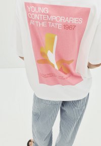 Weißes T-Shirt mit einem pinken Grafikdesign, das den Text "YOUNG CONTEMPORARIES AT THE TATE 1967" und abstrakte Formen in Gelb und Weiß zeigt. Blau gestreifte Hose.