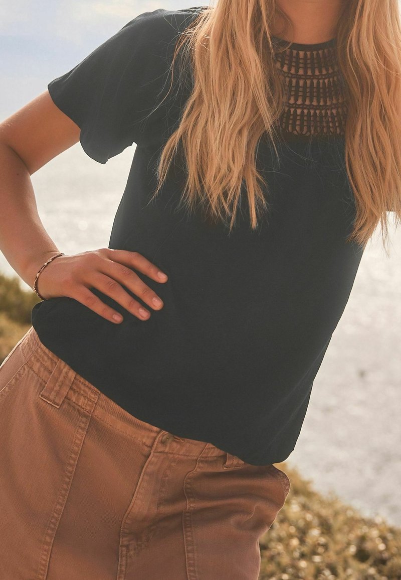 Femme aux longs cheveux blonds portant un haut noir avec un col à motif et un pantalon marron, debout à l'extérieur près de l'eau.
