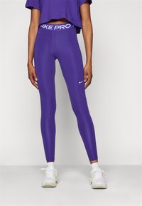 Fioletowe legginsy Nike Pro z szerokim, markowym pasem w talii i małym logo na udzie, w połączeniu z białymi butami sportowymi.